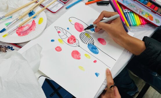 Photo of someone drawing, a hand is visible painting on a colourful page surrounded by arts and crafts tools